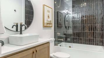 A modern bathroom with a white vanity, a large mirror, a framed picture, a glass shower enclosure, and a white toilet.