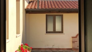 A view through a window of a building with a tiled roof, a small window, and a potted plant with red flowers. - Villa Nuragica - Deluxe Apartment (Nuraxi Figus)