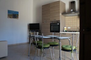 A modern kitchen with a flat-screen TV, a built-in oven, and a dining area with metal chairs and green cushions.