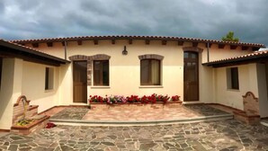 A building with a tiled roof, two windows with shutters, a small porch with stone flooring, and a flower bed with red and pink flowers. - Villa Nuragica - Deluxe Apartment (Nuraxi Figus)