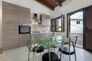 A modern kitchen with a dining area, featuring a glass table and chairs, a built-in oven, and a view of the outside. - Villa Nuragica - Deluxe Apartment (Nuraxi Figus)