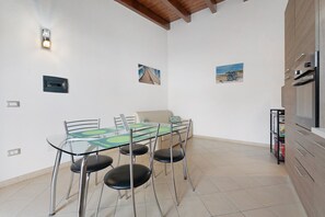 A dining area with a glass table and metal chairs, a sofa, and a kitchen area with cabinets and a microwave. - Villa Nuragica - Deluxe Apartment (Nuraxi Figus)