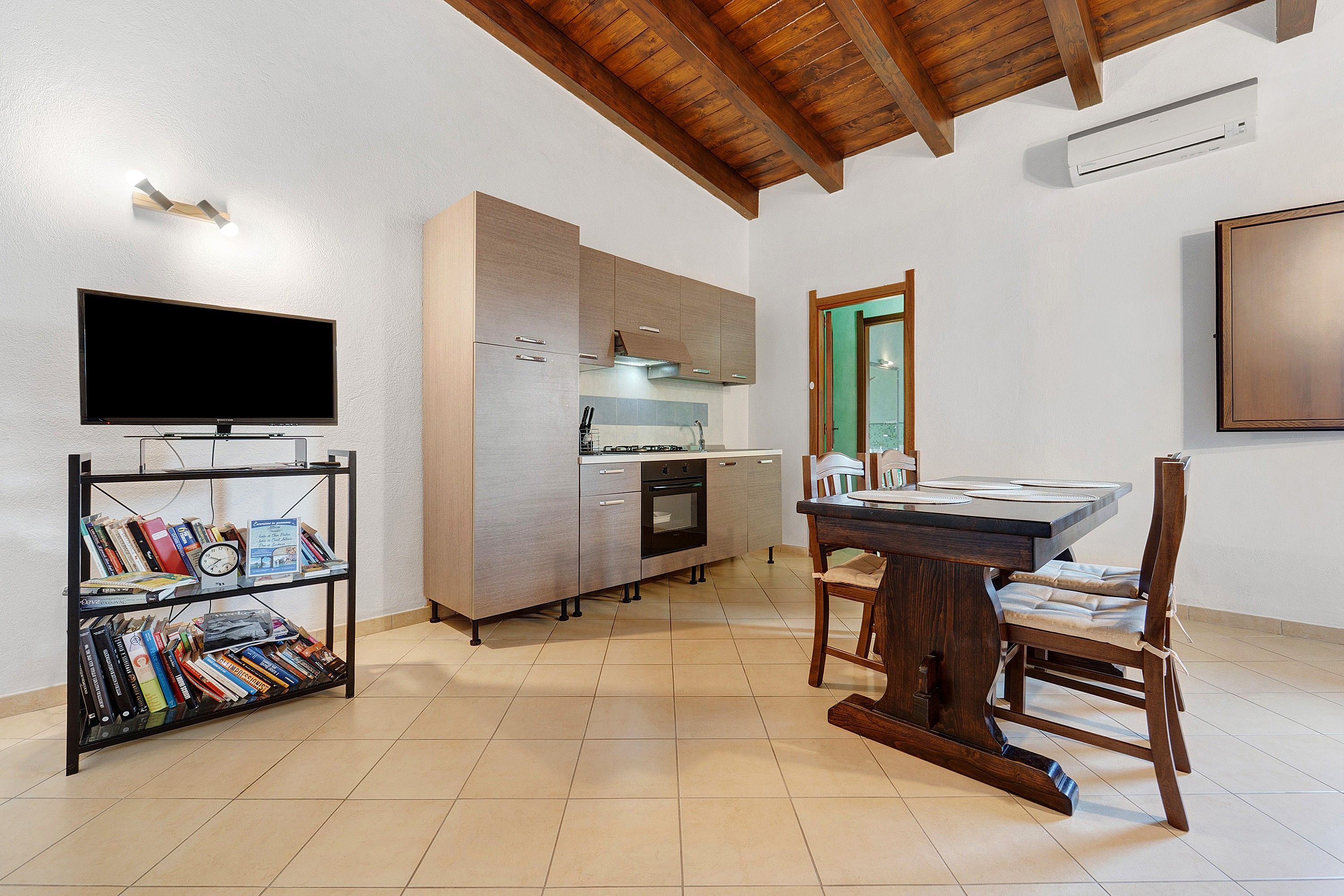 A modern kitchen with a built-in oven, a dining table with chairs, a bookshelf, and a flat-screen TV mounted on the wall.