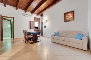 A living room with a dining area, a beige sofa, wooden beams on the ceiling, and a framed picture on the wall. - Villa Nuragica - Classic Apartment (Nuraxi Figus)