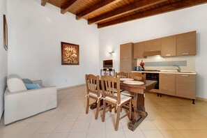A modern kitchen with wooden cabinets, a dining table with chairs, and a sofa in a living area. - Villa Nuragica - Classic Apartment (Nuraxi Figus)