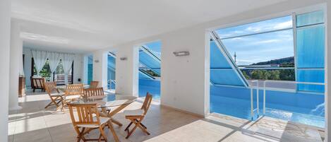 Una habitación amplia con una mesa y sillas de vidrio, vista a una piscina y un balcón con barandilla.
