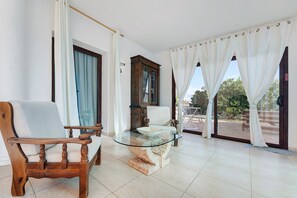 Une pièce comprenant une chaise en bois, une table avec un plateau en verre, un meuble et une vue sur l’extérieur grâce à de grandes fenêtres.