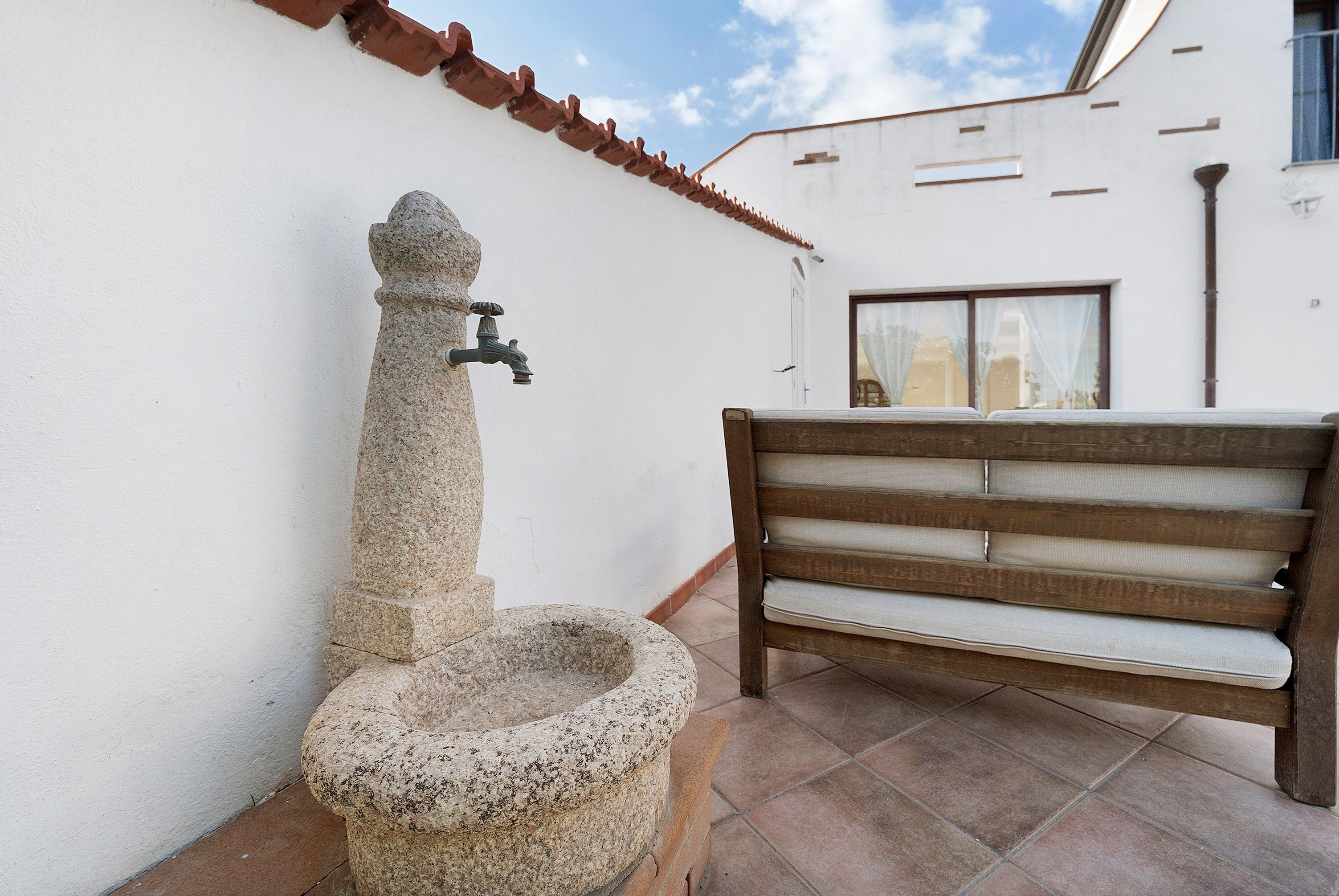 Un bagno esterno in pietra con un rubinetto, una panca di legno e un edificio bianco con tetto in tegole.