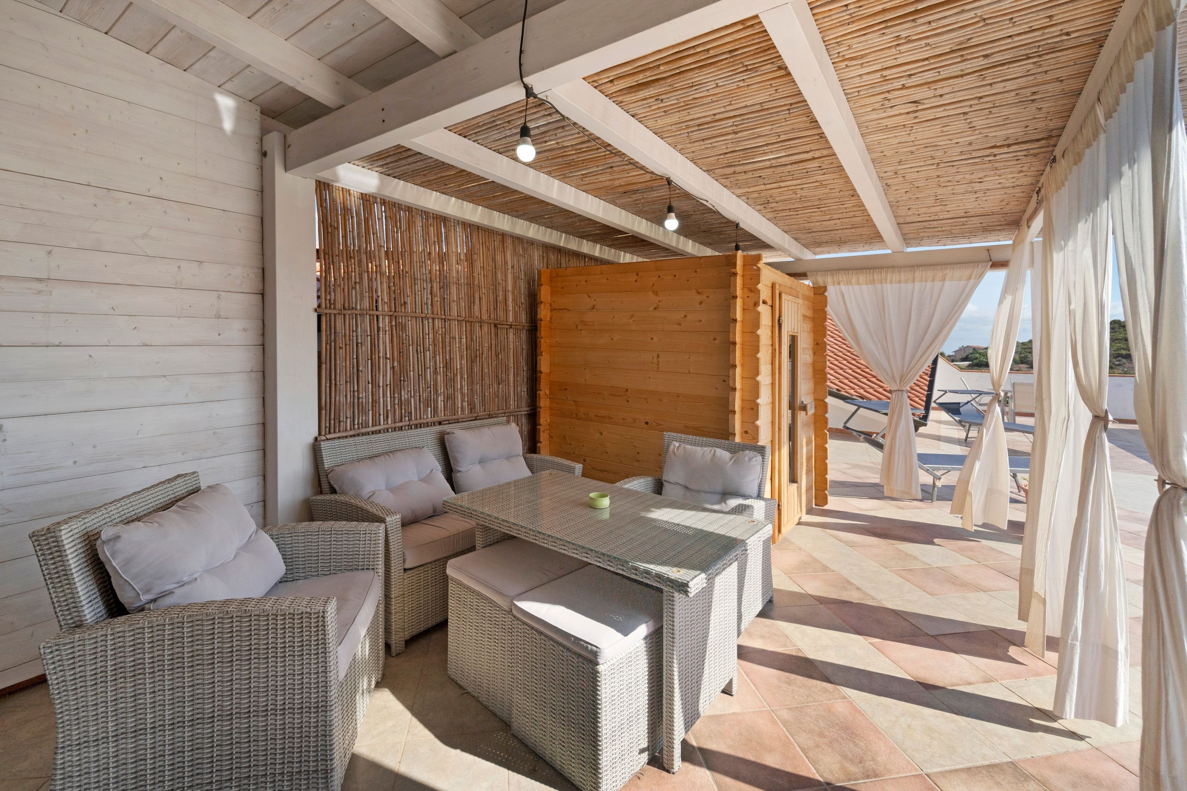 An outdoor seating area with wicker furniture and a glass-top table, a wooden wall, and a view of the beach.