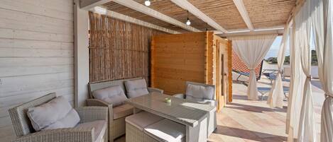 An outdoor seating area with wicker furniture and a glass-top table, a wooden wall, and a view of the beach.