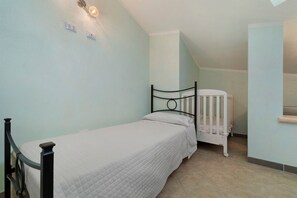 Une petite chambre avec un lit simple, un berceau et un luminaire fixé au mur.
