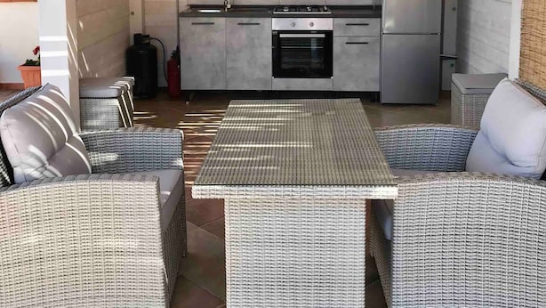 A modern kitchen with grey cabinets, a stainless steel refrigerator, and a dining area with wicker furniture.