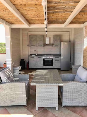 A modern kitchen with grey cabinets, a stainless steel refrigerator, and a dining area with wicker furniture. - Villa Nuragica – Apartment with Outdoor Kitchen and Terrace with View (Nuraxi Figus)