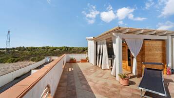 A rooftop terrace with a pergola, potted plants, a sun lounger, and a view of the surrounding landscape.