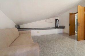 Una habitación con un sofá color beige, un banco estampado, piso de baldosas y una puerta de madera.