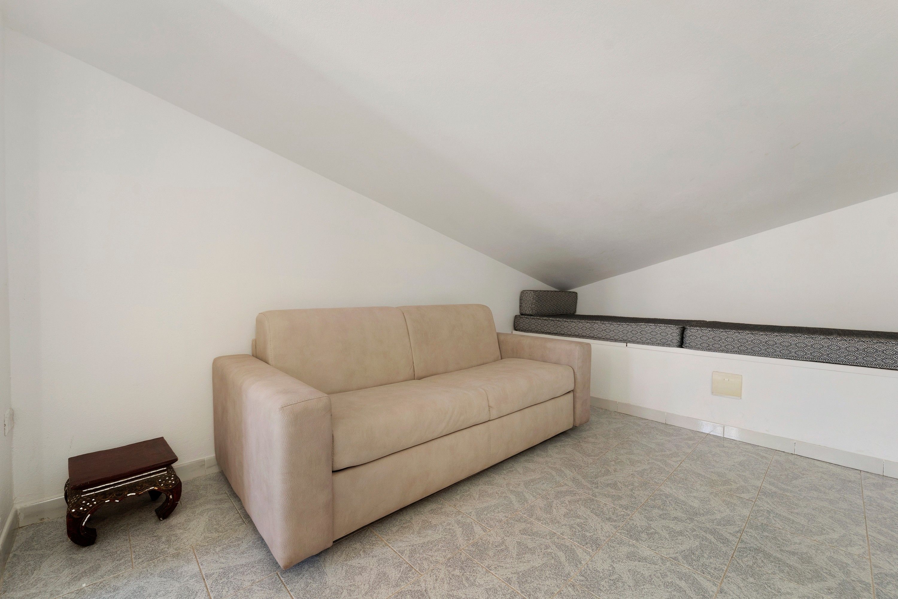 A beige sofa in a corner of a room with a sloped ceiling and a patterned shelf against the wall.