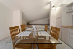 Een eethoek met een tafel met glazen blad en houten stoelen, een keuken met aanrecht en kasten, en een schuin dak.