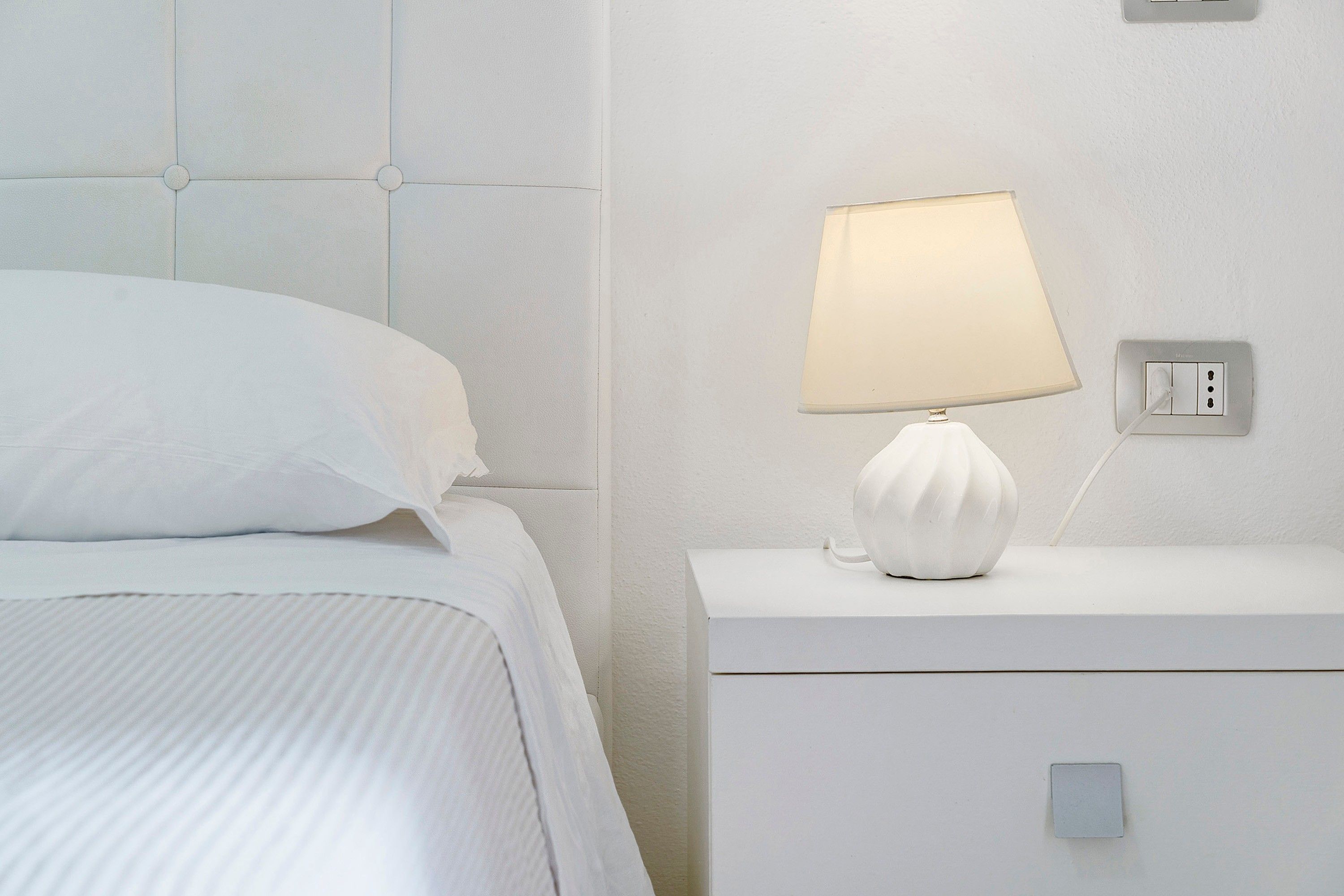 Een net opgemaakt bed met een wit hoofdbord en een nachtkastje met een lamp.