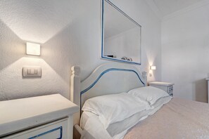 A bedroom with a bed, bedside table, nightstand, and a wall-mounted mirror. - Villa Nuragica – Room with Private Bathroom (Nuraxi Figus)