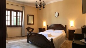 A bedroom with a bed, bedside tables, a chandelier, a window with curtains, and a wall clock. - Villa Nuragica – Room with Private Bathroom (Nuraxi Figus)