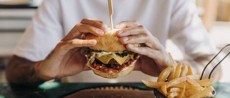 Een persoon houdt een hamburger vast, met een cocktailprikker erin. Op de tafel staat een mand met friet.