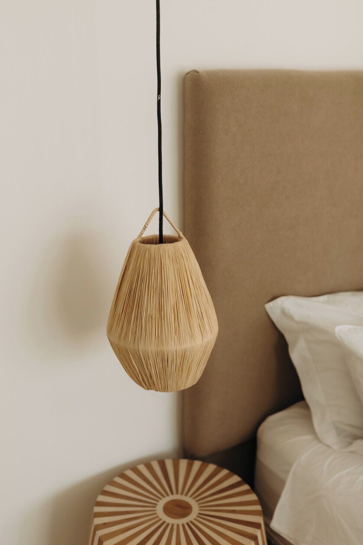 Un luminaire suspendu, fait de fibres tressées, dans une pièce où se trouve un lit et un objet décoratif rond posé sur le sol.