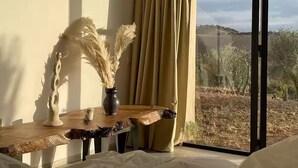 A room with a wooden table, a vase with pampas grass, and a view of a dry landscape through a large window with curtains. - Laho Lodge (Lalla Takarkoust)