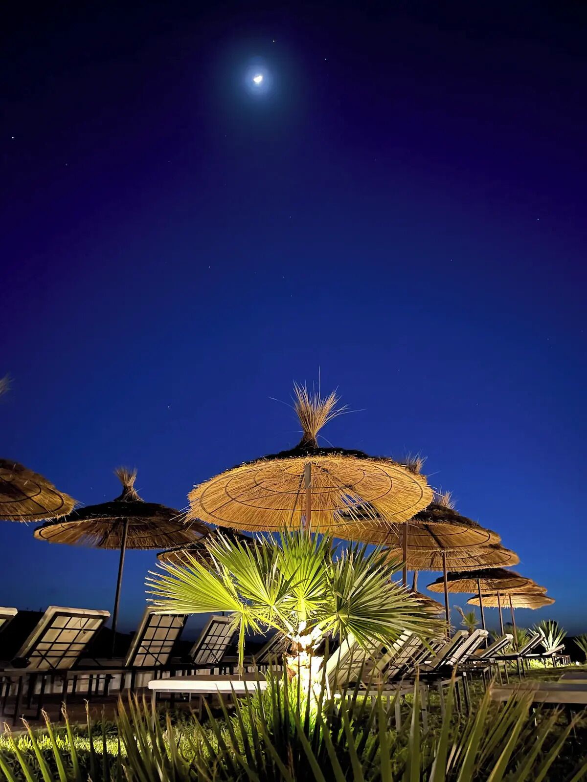 A night scene with thatched umbrellas and lounge chairs, suggesting a resort setting.