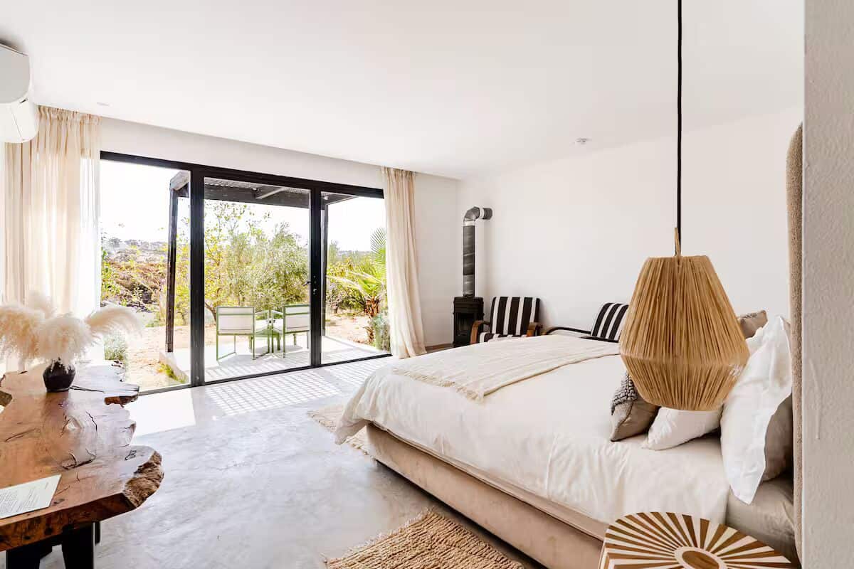 A bedroom with a large bed, a wooden table, a hanging lamp, and a view of the outdoors.