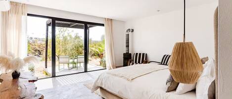 A bedroom with a large bed, a wooden table, a hanging lamp, and a view of the outdoors.
