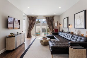 A modern living room with a sectional sofa, a rattan console, and a large window with curtains. - Luxury 4BR | Near Swedish | Sauna + Hot Tub (Englewood)