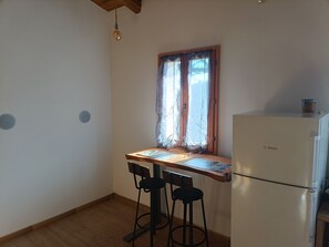 A small kitchen with a wooden table, two black bar stools, a white Bosch refrigerator, and a window with sheer curtains. - The house on the rainbow (Piemonte)