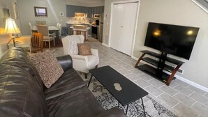 A living room with a dark leather sofa, a white armchair, a black coffee table, and a flat-screen TV on a wooden stand.
