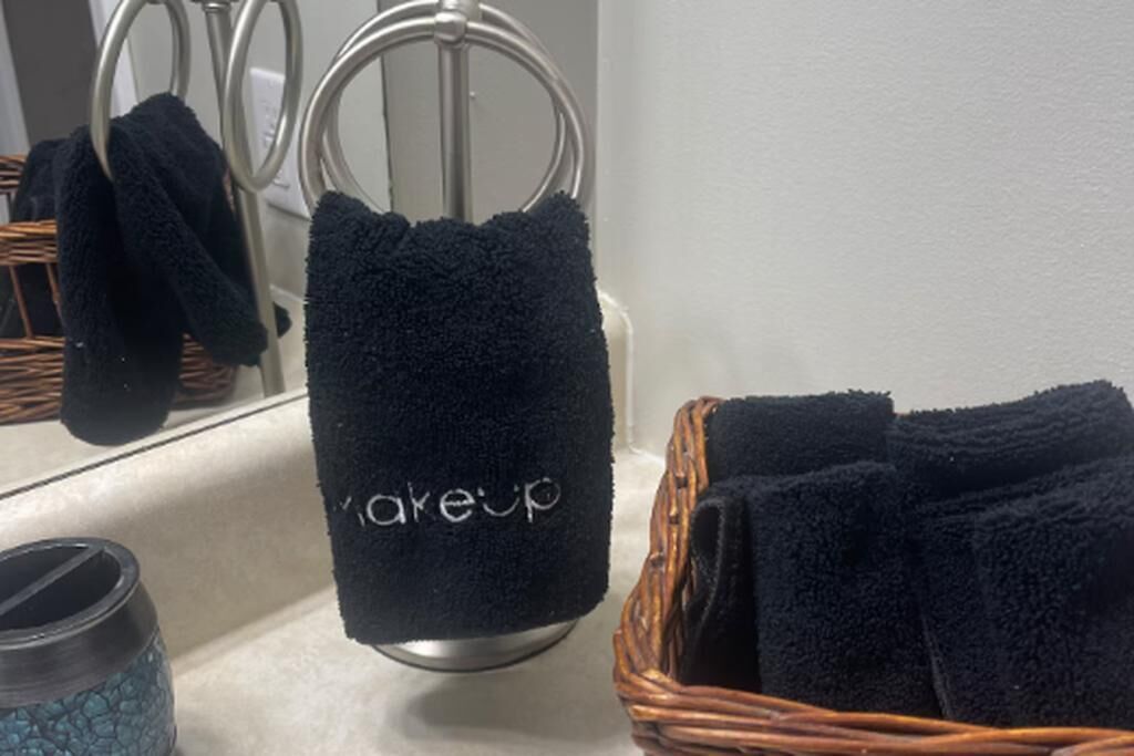 A bathroom with a towel rack holding a black towel with "makeup" written on it, a wicker basket with towels, and a blue textured container.