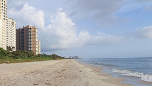 A sandy beach with footprints leading to the ocean, tall buildings in the background, and a clear sky. - Regatta at Vanderbilt Beach | Pool | Walk to Gulf (Naples)