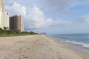 Ein sandiger Strand mit Fußspuren, die zum Meer führen, hohe Gebäude im Hintergrund und ein klarer Himmel.