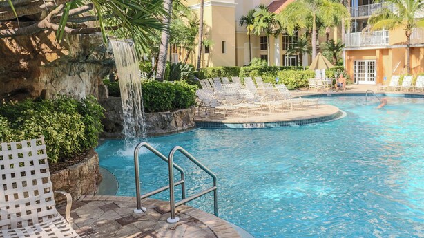 A pool area with a waterfall feature, lounge chairs, and a stone wall. - Regatta at Vanderbilt Beach | Pool | Walk to Gulf (Naples)