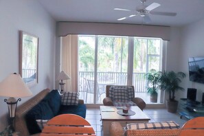 Ein Wohnzimmer mit einer Couch, Sesseln, einem Couchtisch und einem Fernseher. Es gibt einen Balkon mit Stühlen und einem Tisch.