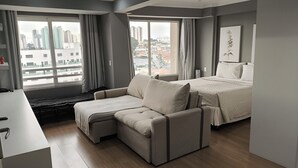 Een moderne hotelkamer met een bank, een bed, een bureau en grote ramen met uitzicht op de stad.