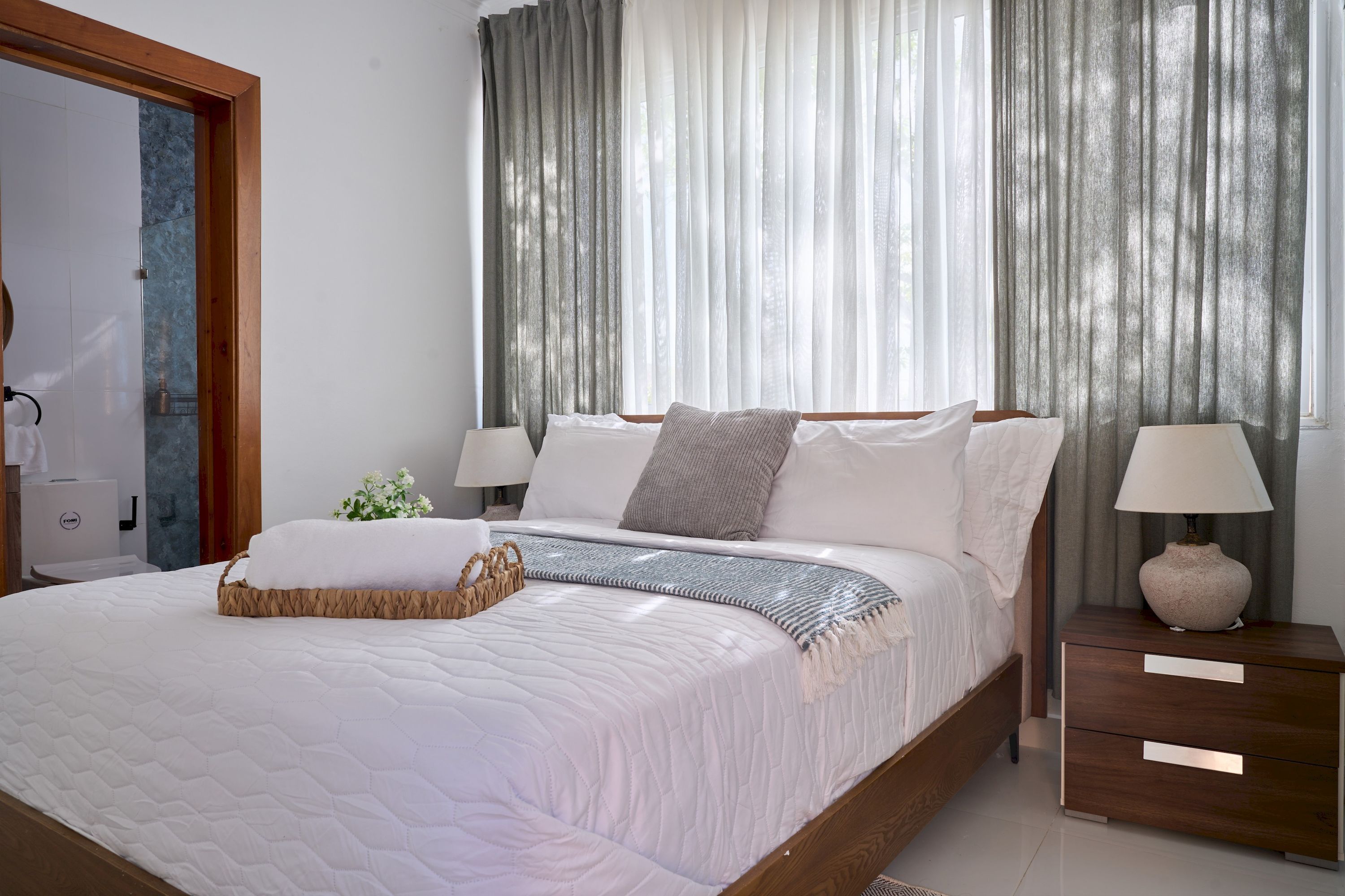 A neatly made bed with a white quilt, a bedside table with a lamp, and a wooden nightstand.