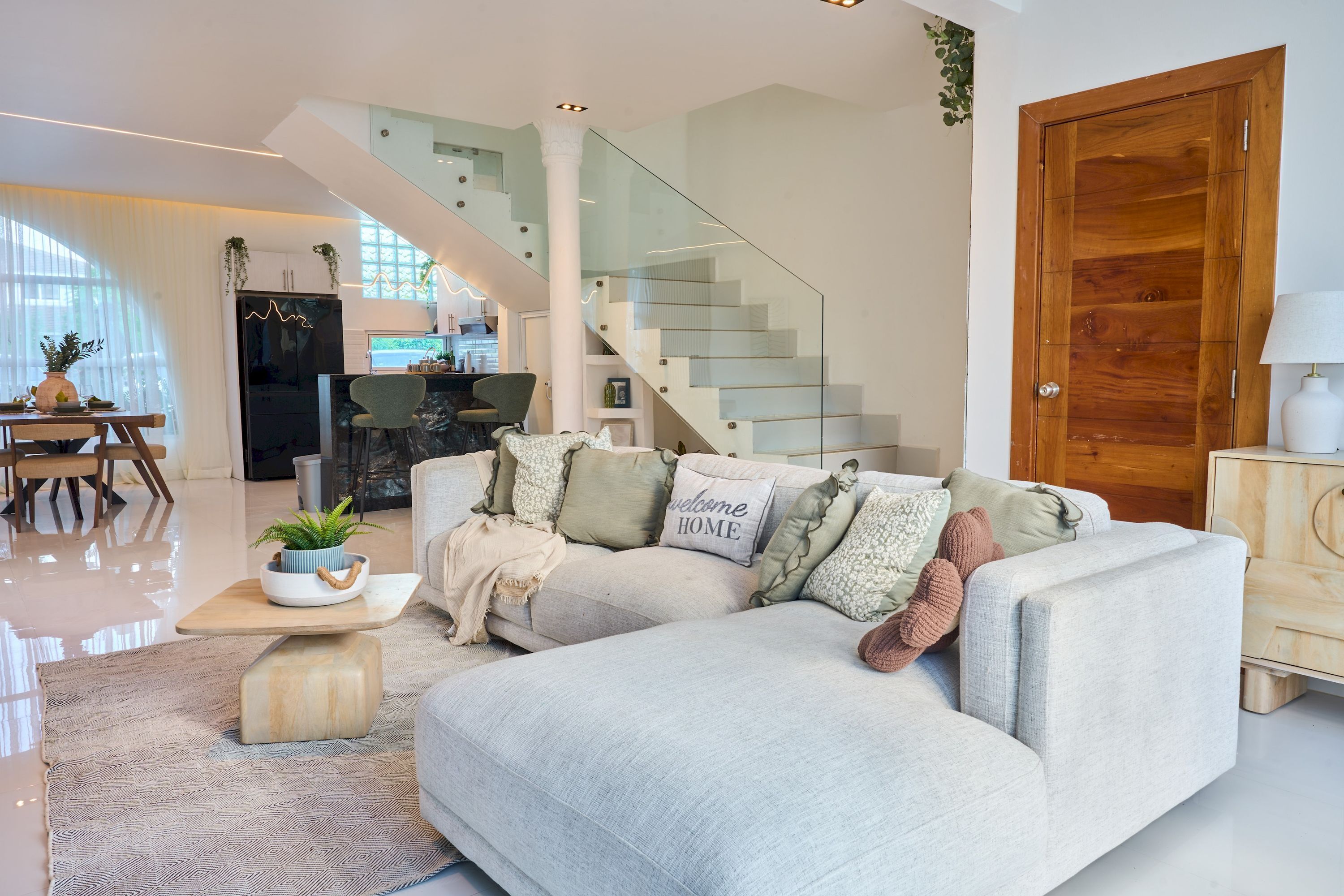 A modern living room with a light-colored sectional sofa, a glass staircase, and a wooden door.