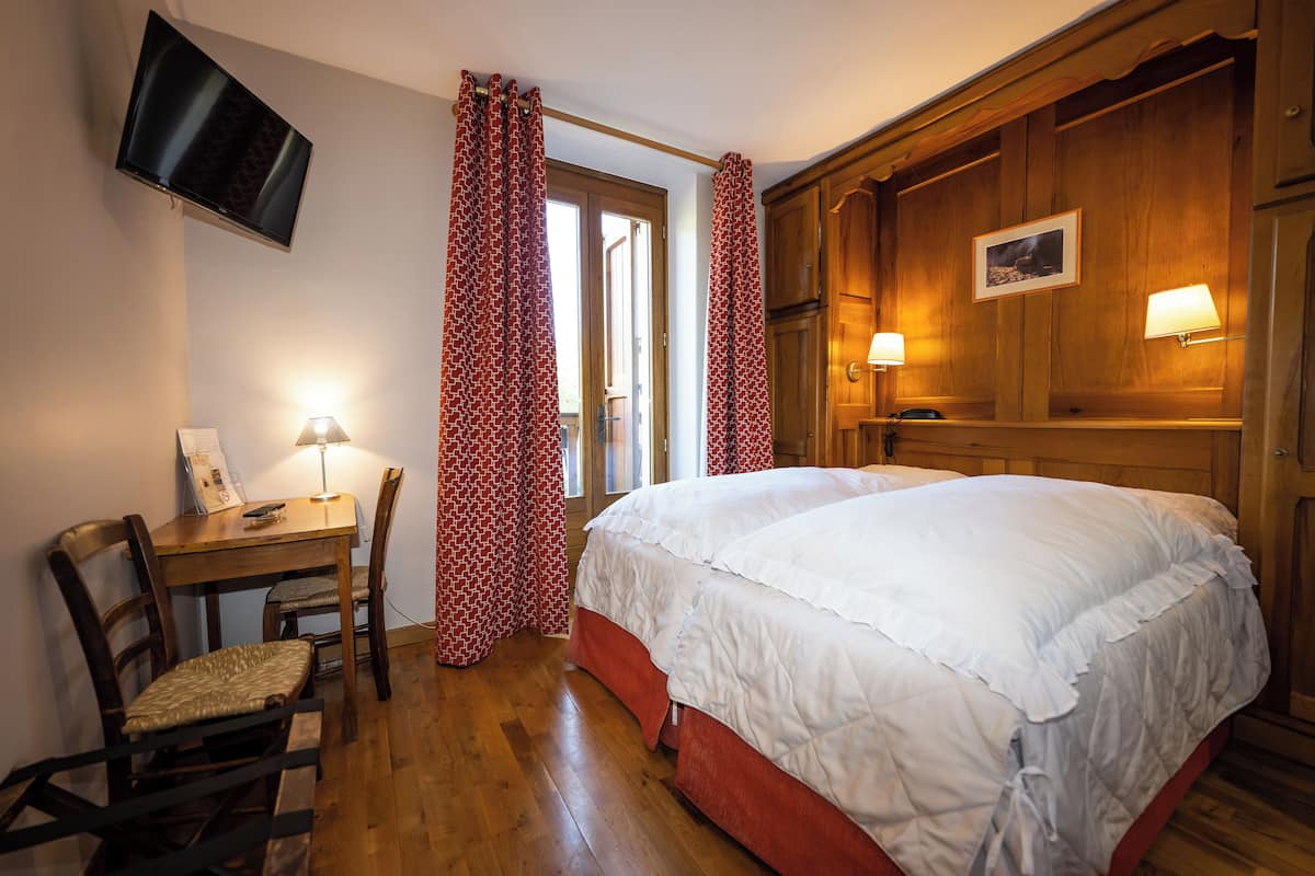 Ein Schlafzimmer mit Bett, Schreibtisch mit Stuhl, einem an der Wand befestigten Fernseher und einer Tür mit Vorhängen.