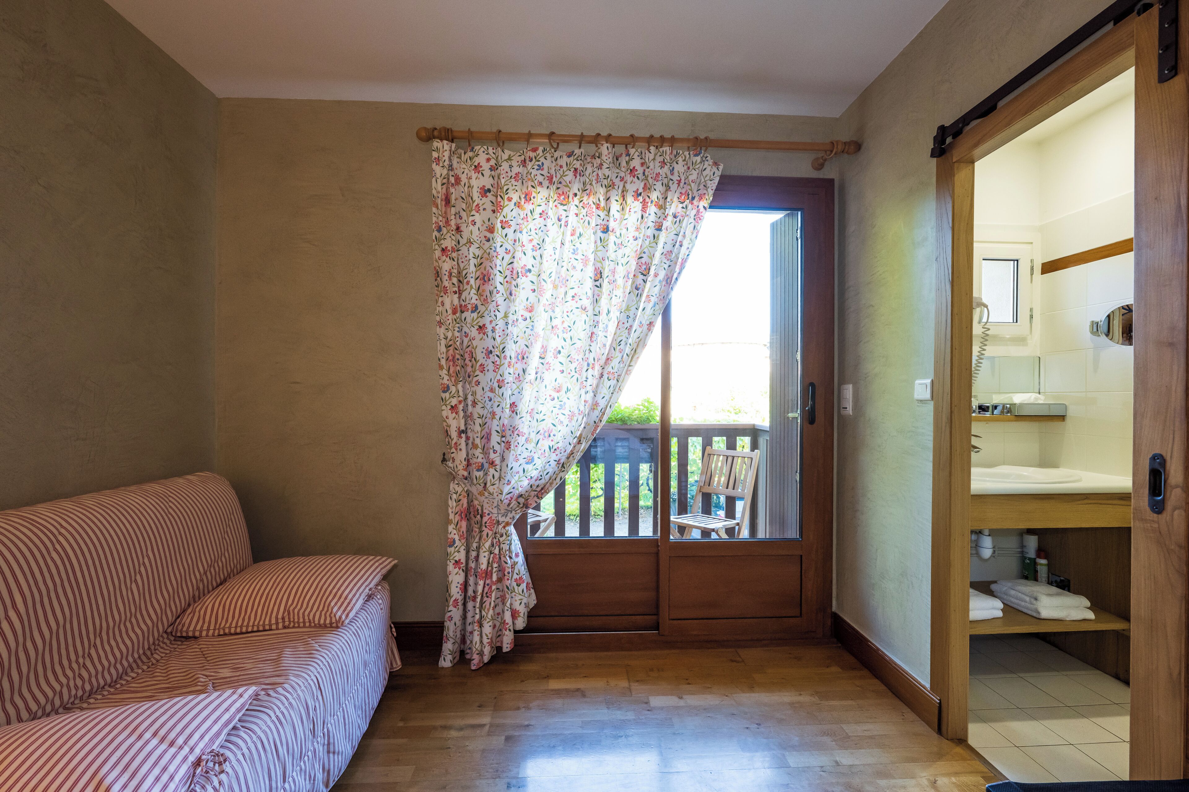 A room with a wooden floor, a striped sofa, a wooden door leading to a balcony, and a bathroom with a wooden door.