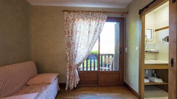 A room with a wooden floor, a striped sofa, a wooden door leading to a balcony, and a bathroom with a wooden door.