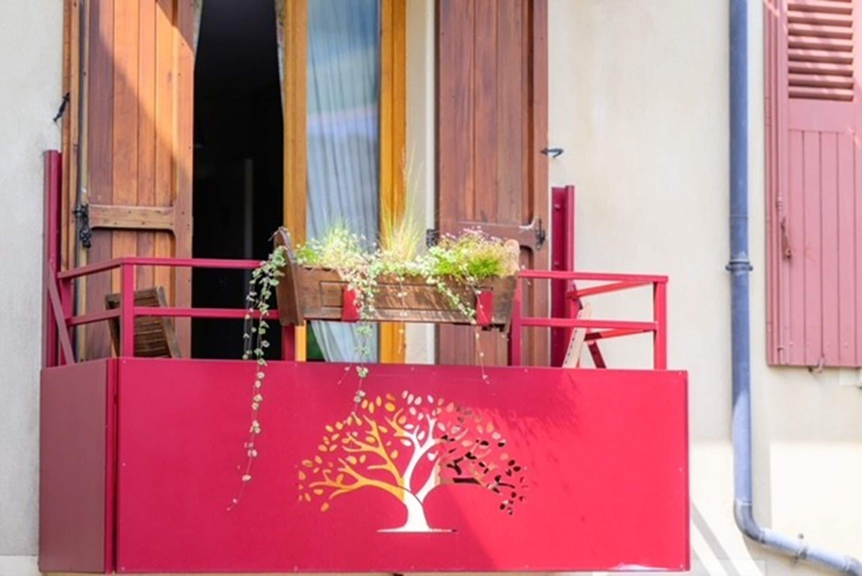 Un balcón con barandilla roja, una jardinera de madera con plantas y una caja roja con diseño de árbol.