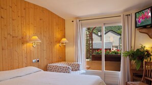 Habitación de hotel con pared de madera, cama, sillón, mesa, televisión y balcón con flores.