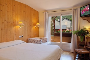 Quarto de hotel com parede de madeira, uma cama, uma cadeira, uma mesa, uma televisão e uma varanda com flores.