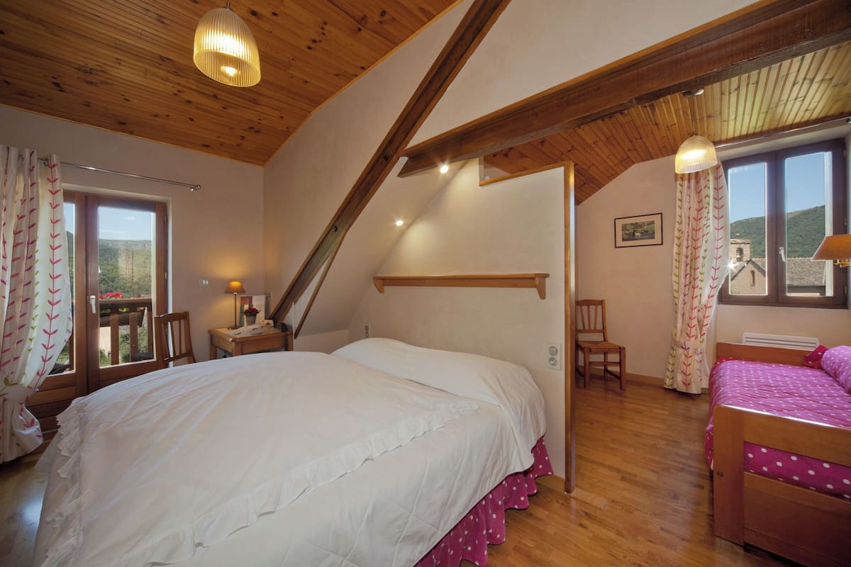 Ein Schlafzimmer mit einem großen Bett, einem kleineren Bett, Holzbalken, einem Fenster und einem Stuhl.