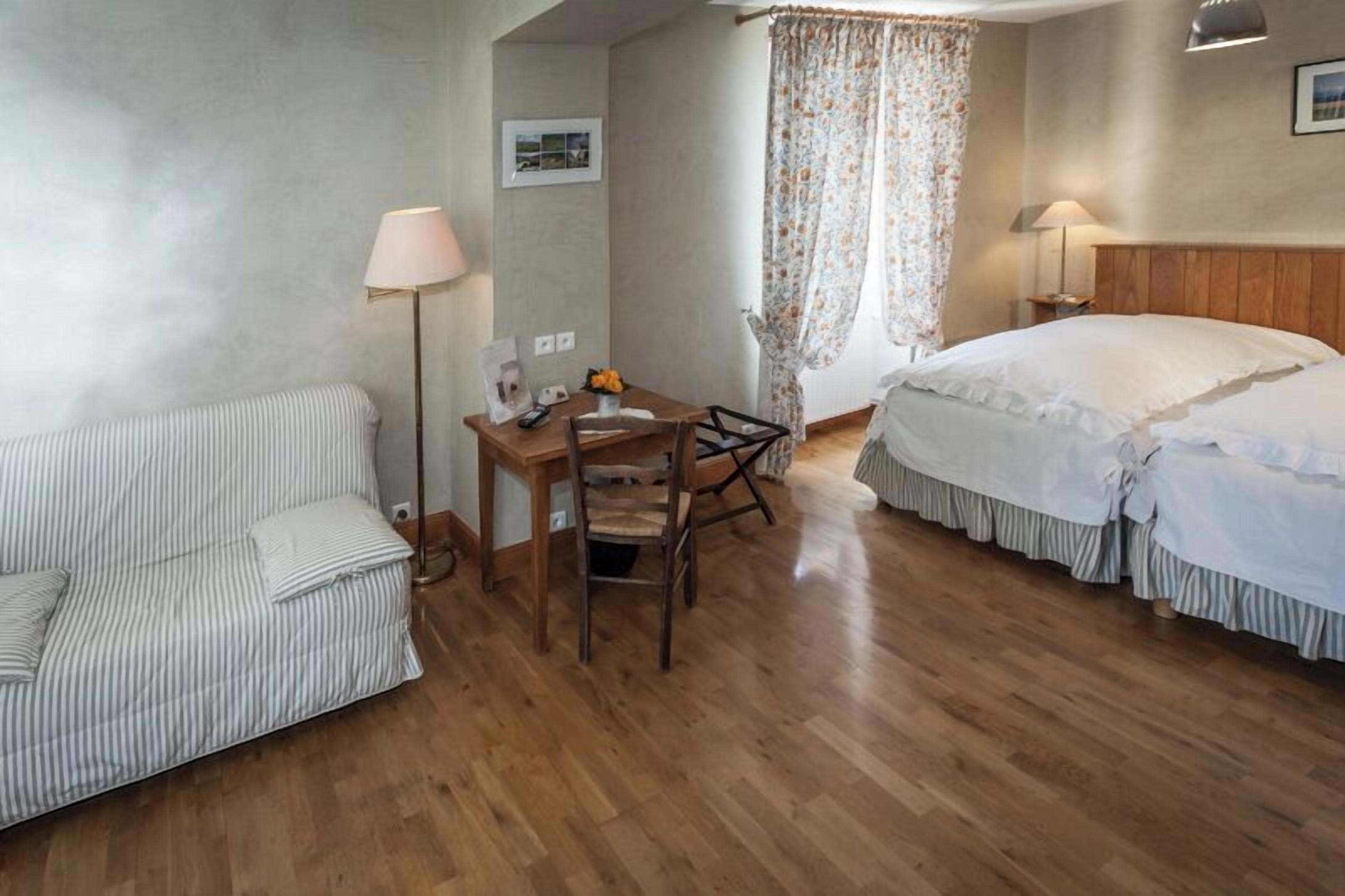 Quarto de hotel com cama, sofá, escrivaninha com cadeira, luminária e janela com cortinas.
