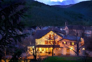 A stone building with a balcony, surrounded by trees and hills. - Hôtel Restaurant la Lozerette (Bédouès-Cocurès)